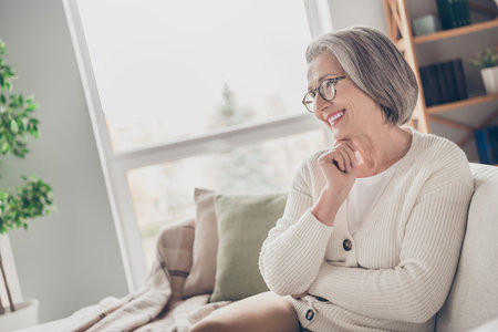 Photo of pretty adorable senior lady dressed white cardigan sitting sofa watching movie indoors house roomの写真素材