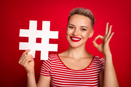 Photo of cheerful satisfied girl wear stylish striped clothes hold paper card ad okey symbol feedback isolated on red color backgroundの写真素材