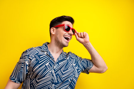 Profile portrait of impressed positive young man toothy smile arm touch sunglass empty space isolated on yellow color backgroundの写真素材