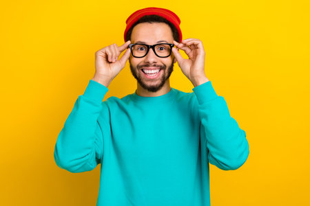 Portrait of satisfied friendly man with beard dressed turquoise pullover fingers touching glasses isolated on yellow color backgroundの写真素材