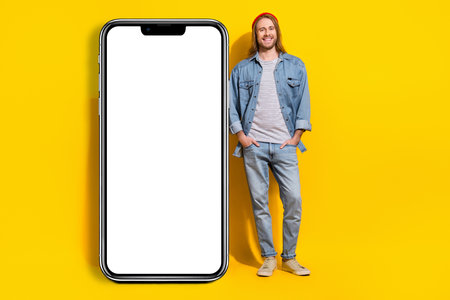 Full size photo of good mood man jacket jeans trousers standing near board presenting smartphone...
