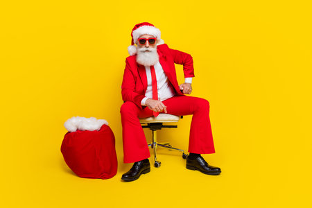 Full size portrait of confident serious aged man sit chair big presents bag isolated on yellow color backgroundの写真素材