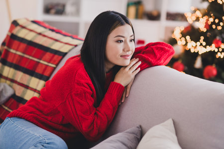 Photo of pretty optimistic positive girl with long hairstyle dressed red knit sweater laying on couch look empty space in house indoorsの写真素材
