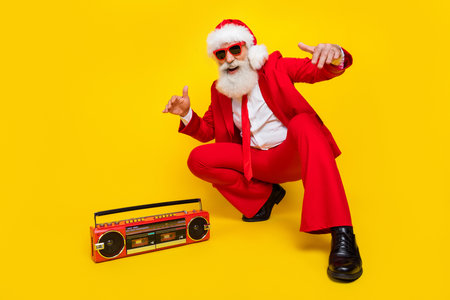 Full length photo of funky cool man pensioner dressed red suit santa hat listening boom box songs isolated yellow color backgroundの写真素材