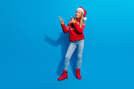Full length photo of adorable teen girl wear jumper santa hat directing look at offer empty space isolated on blue color backgroundの写真素材
