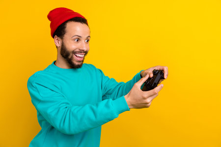 Photo of impressed guy wear teal sweatshirt red beanie hat look empty space hold game console joystick isolated on yellow color backgroundの写真素材