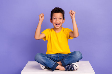 Full length photo of nice young pupil boy raise fists lucky sit white cube dressed yellow garment isolated on violet color backgroundの写真素材