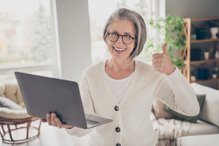 Photo of smiling confident elderly lady wear white showing thumb up texting modern gadget indoors...