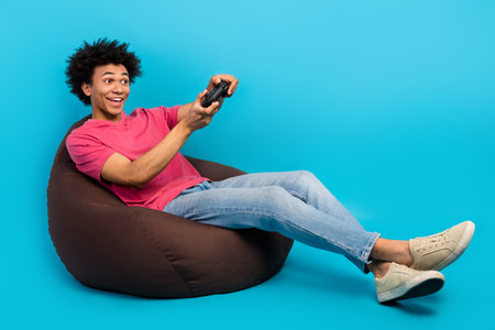 Full length photo of nice young guy sit brown beanbag excited play joystick wear trendy pink outfit...