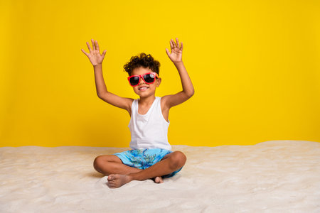 Full length photo of sweet cheerful small boy wear white shirt dark eyewear siting beach rising arms empty space isolated yellow color backgroundの写真素材