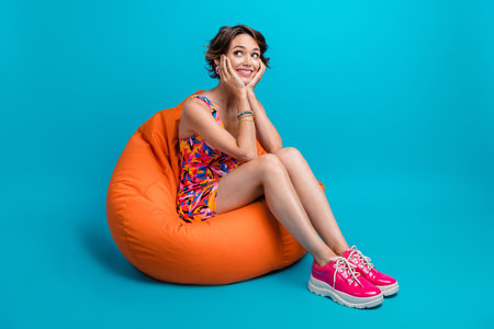 Full length photo of adorable thoughtful lady wear print clothes arms cheeks sitting bean bag...