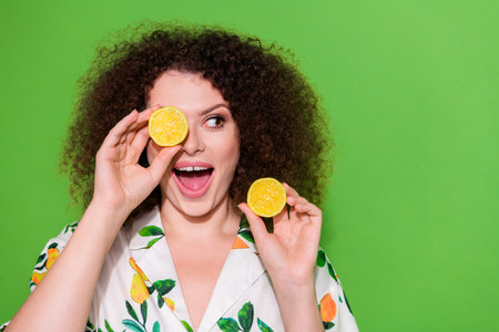 Portrait of astonished funky lady hold lemon halves cover one eye look empty space isolated on green...