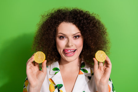 Photo of gorgeous cheerful lady tongue lick teeth hold two halves fresh lemons isolated on green...