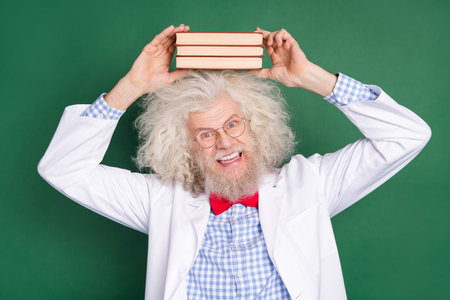 Photo of funky funny cheerful smiling good mood crazy scientist hold books on head isolated on green color backgroundの写真素材