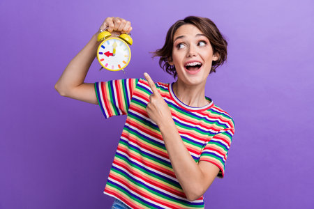 Photo of glad cute girl dressed stylish striped clothes looking alarm clock isolated on purple color...