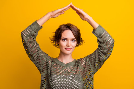 Photo of charming cute girl dressed stylish clothes shows roof above her head dream buy house isolated on yellow color backgroundの写真素材