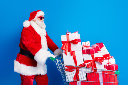 Photo portrait of mature pensioner man push supermarket cart wear trendy santa claus costume coat isolated on blue color backgroundの写真素材