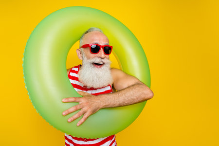 Photo of carefree excited senior man wear red striped swimsuit holding inflatable circle looking empty space isolated yellow color backgroundの写真素材