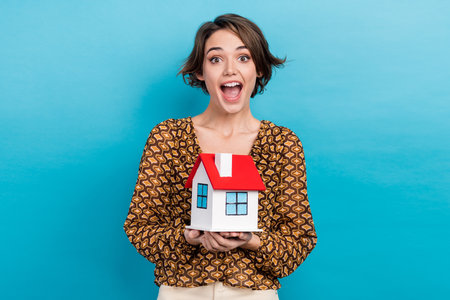 Photo of young funny reaction girl holding her first ownership building apartment open mouth speechless isolated on blue color backgroundの写真素材