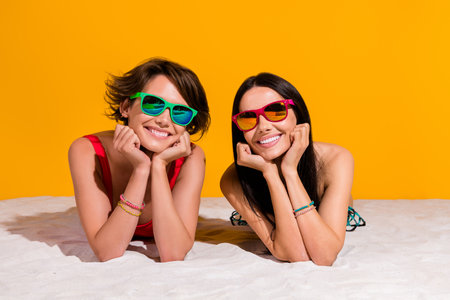 Photo of sexy cheerful girls dressed swimsuits dark eyewear lying sand isolated yellow color backgroundの写真素材