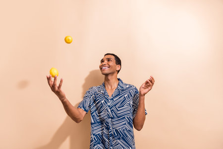 Photo of funny cheerful guy dressed print shirt smiling juggling lemons isolated beige color backgroundの写真素材