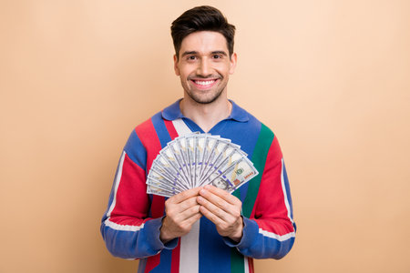 Photo of nice positive young man toothy smile arms hold dollar bills isolated on beige color backgroundの写真素材