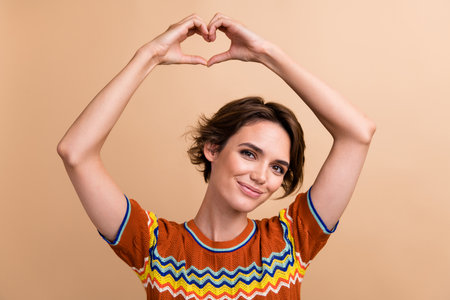 Portrait of stunning lovely young lady hands show heart symbol above head isolated on beige color...
