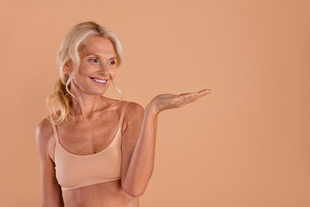 Photo of cute dreamy senior lady dressed lingerie holding arm looking empty space isolated beige color backgroundの写真素材