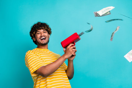Photo of overjoyed successful guy hold money gun shooting flying dollar bills isolated on teal color backgroundの写真素材