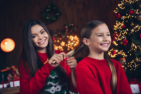Photo of sweet shiny small sisters wear red jumpers brushing hair making xmas hairstyle indoors new year north pole houseの写真素材