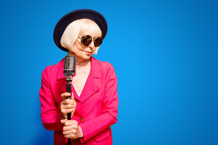 Portrait of grandmother in a hat and red jacket holds a microphone stand and looks sideways isolated on brigth yellow background with copy spaceの写真素材