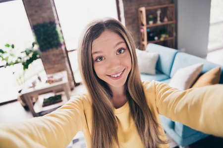 Selfie portrait of cheerful young teenager smiling girl wearing yellow pullover showing her new living room in parents apartments indoorsの写真素材