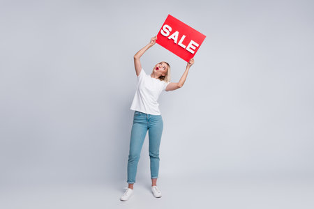 Full length photo of funky astonished girl wear stylish t-shirt looking at sale placard over head isolated on gray color backgroundの写真素材