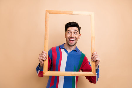 Photo of smiling funky guy holding wooden portrait frame looking empty space laughter sale...