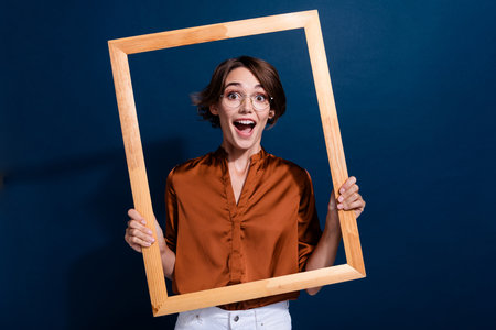 Photo portrait of lovely young lady hold wooden frame excited photo dressed stylish brown blouse isolated on dark blouse color backgroundの写真素材