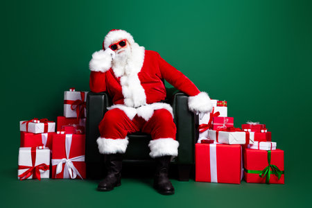 Full length photo of cute funny claus wear red costume preparing christmas gifts sitting armchair isolated green color backgroundの写真素材