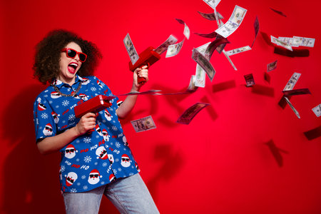 Photo of lucky shocked woman wear ugly santa claus print x-mas shirt shooting cash flying gun...