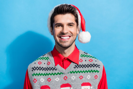 Photo portrait of handsome young guy toothy beaming smile wear x-mas print vest hat outfit isolated on blue color backgroundの写真素材