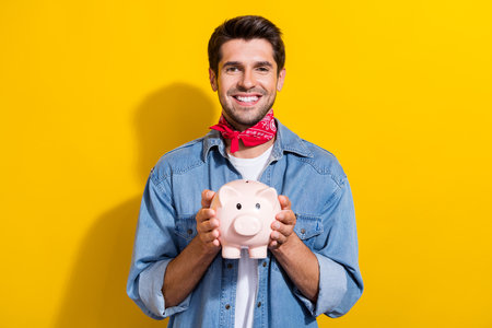 Photo portrait of nice young man hold piggy account toothy smile wear trendy jeans garment isolated on yellow color backgroundの写真素材
