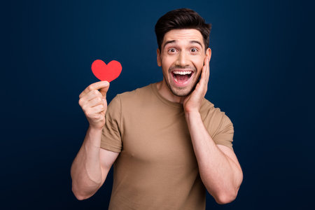 Photo of astonished crazy man dressed beige t-shirt touching cheekbone hold red heart postcard isolated on dark blue color backgroundの写真素材