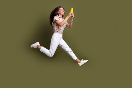 Full length photo of blogging lady escaping home with smartphone jumping holding smartphone writing blog isolated on khaki color backgroundの写真素材