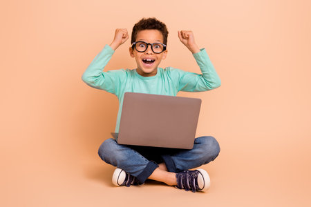 Full body portrait of small delighted schoolchild sit floor use netbook raise fists isolated on...