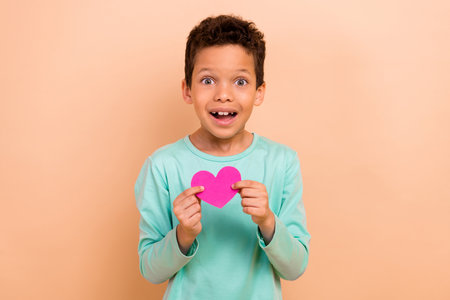 Photo of charming cheerful boy wear trendy turquoise clothes hands hold heart shape paper card isolated on beige color backgroundの写真素材