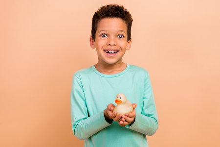 Photo of cheerful carefree playful boy wear trendy turquoise clothes hands hold yellow duck isolated on beige color backgroundの写真素材