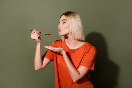 Photo of young girl dreaming to taste salt in her soup wearing orange t shirt making homemade recipe isolated on khaki color backgroundの写真素材