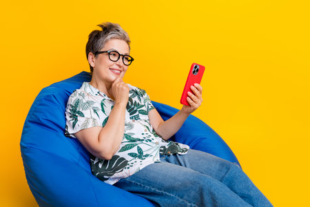 Photo portrait of attractive senior woman sit bean bag video call wear trendy tropical print clothes isolated on yellow color backgroundの写真素材