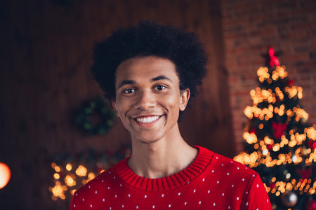 Photo of positive cheerful guy wear red ugly xmas sweater smiling celebrating christmas indoors apartment roomの写真素材