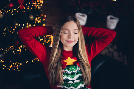 Photo of peaceful relaxed schoolgirl with blond hair wear red sweater eyes closed palms behind head on christmas eve at home indoorsの写真素材