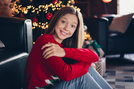 Photo of toothy beaming schoolgirl with blond hairstyle wear red sweater sit on floor hug herself at christmas morning at home indoorsの写真素材