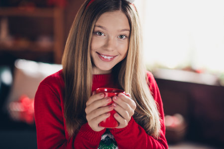 Closeup photo of cute smiling teenage girl youngster holding red cup tea or eggnog enjoying christmas vibe cold weather atmosphere at homeの写真素材